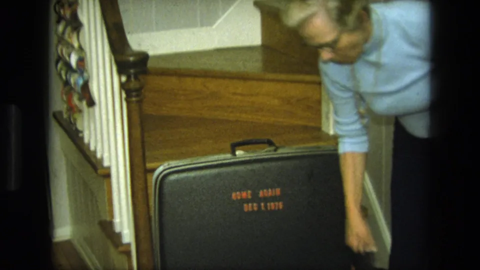 In a 1975 California home interior, a blond woman in glasses bends near a black suitcase by a wooden staircase.