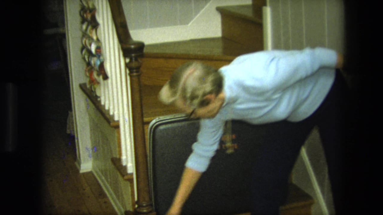 Woman leans forward toward a black suitcase near the stair rail inside a home.