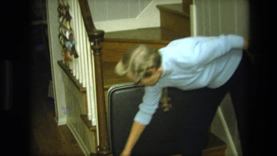 The woman leans across the lower stairs toward a black suitcase in a private California home interior.