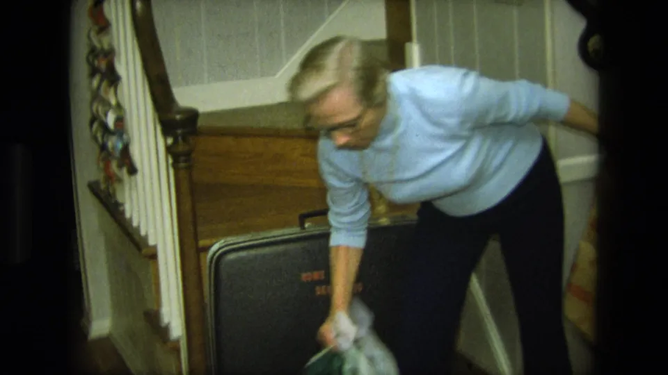 A blond woman in a blue sweater holds a light item while standing over a black suitcase near the stairs.