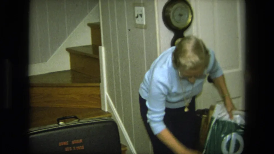 The woman bends near a black suitcase and soft household or travel items at the bottom of a wooden staircase.