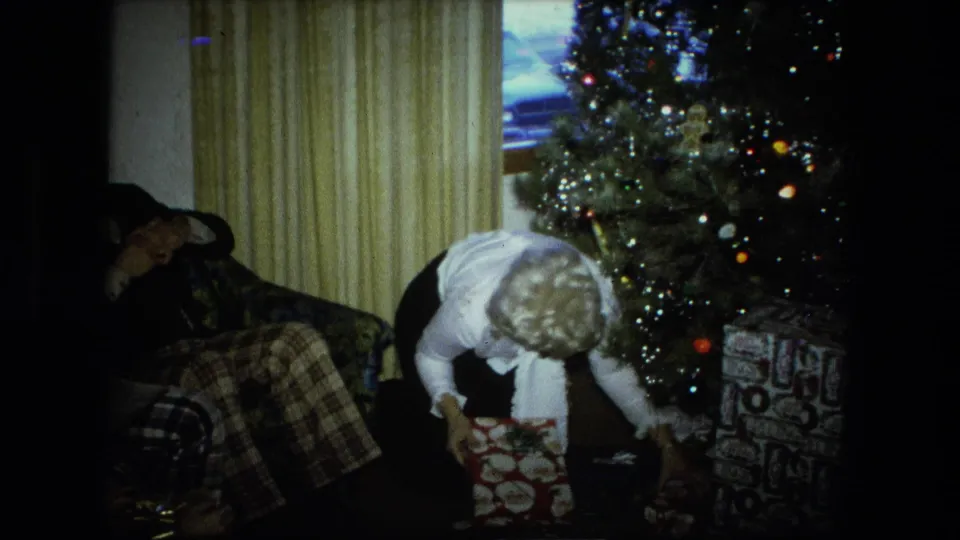 In a 1976 Fort Wayne home-movie frame, a blonde woman in a white blouse bends to unwrap gifts beside a lit Christmas tree.
