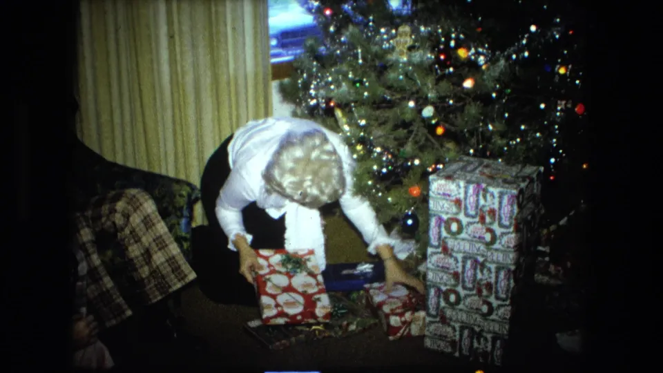 A 1976 Fort Wayne living room frame shows a woman kneeling near presents beneath a decorated and lit Christmas tree.