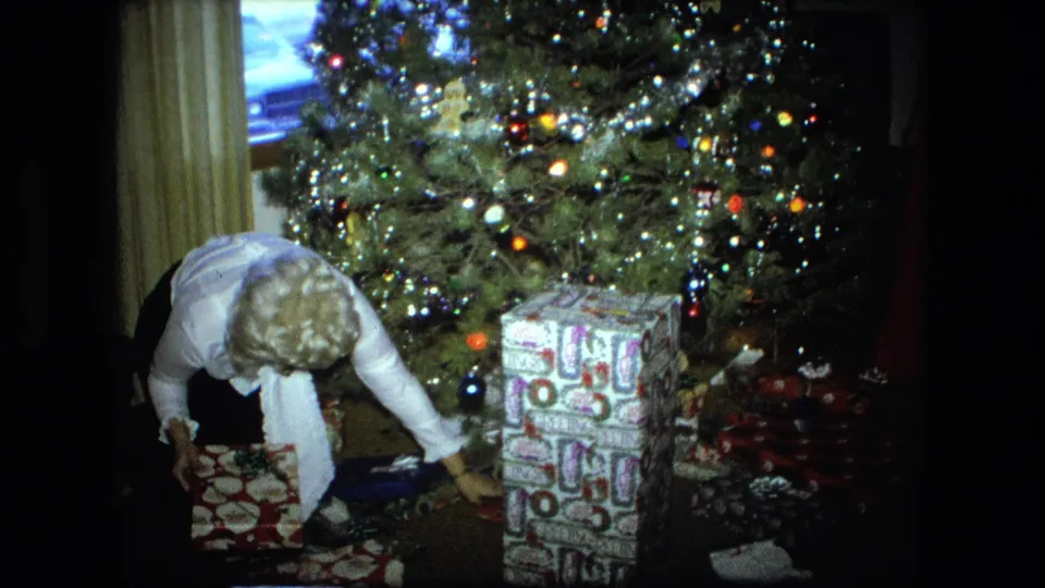 The 1976 Fort Wayne Christmas scene continues as the woman reaches across wrapped gifts near a lit tree and stacked boxes.