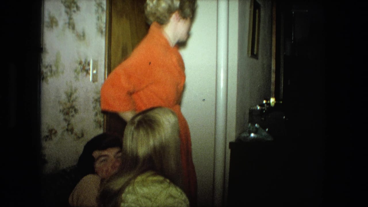Woman in orange sweater stands in a hallway while seated people are partly visible nearby.