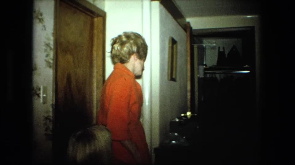 A woman in an orange sweater passes through a dim Fort Wayne home hallway with wood doors, wall decor, and an open closet visible.
