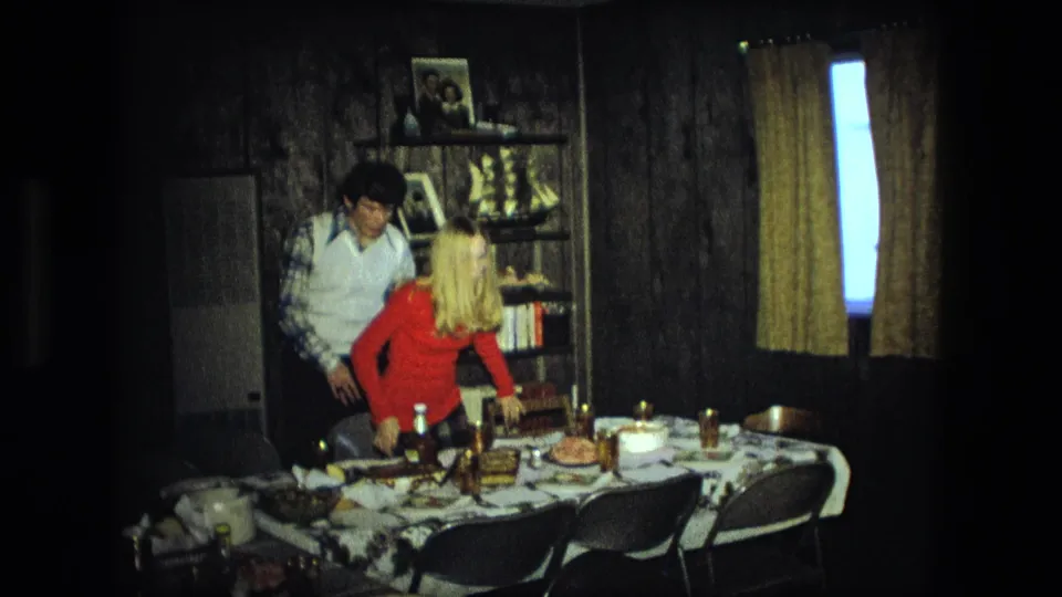 Adults in a 1976 Fort Wayne home stand around a table set with food, bottles, cups, and folding chairs in a wood-paneled room.