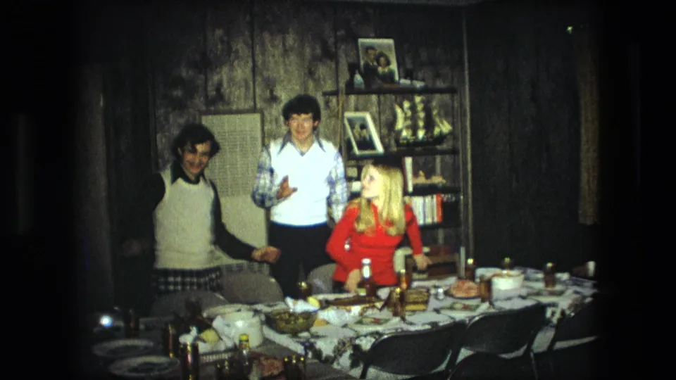 Three adults gather around a set table in a Fort Wayne home, with shelves, framed photos, curtains, and wood paneling behind them.