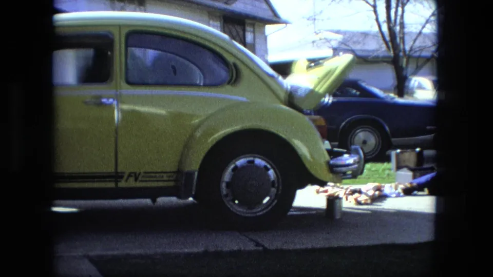 1976 Fort Wayne home-movie frame showing a yellow Volkswagen Beetle with its hood open, tools nearby, and other cars parked in a residential driveway.