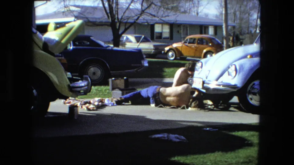 1976 Fort Wayne home-movie frame showing a man lying on a driveway near tools and parts, with Volkswagen Beetles and a blue sedan around him.