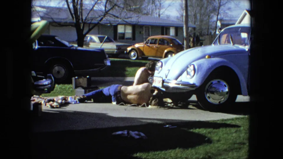 1976 Fort Wayne frame showing a man on the driveway near a pale blue Volkswagen Beetle, with tools, parts, a blue sedan, and an orange Beetle visible.