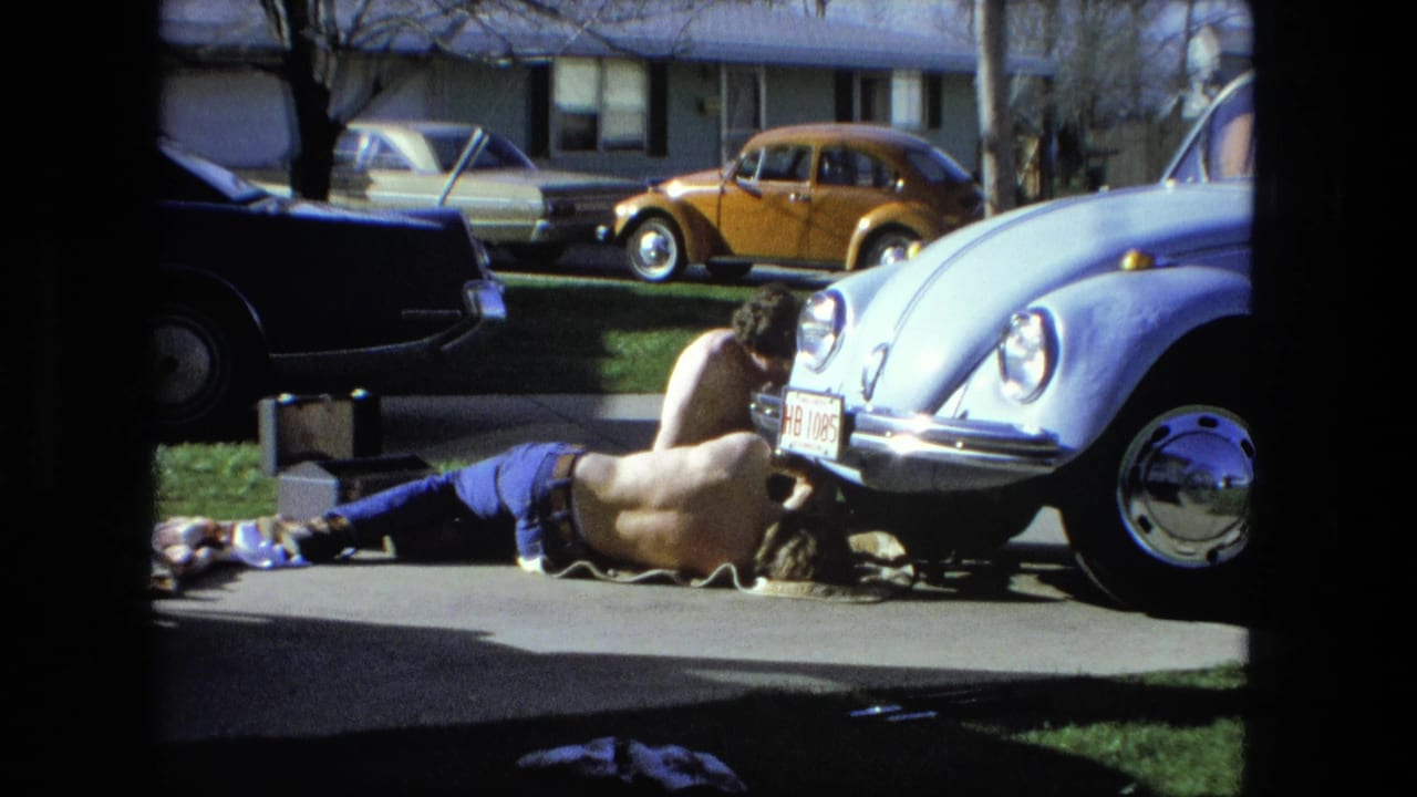 Two shirtless men work near the front of a light blue Volkswagen Beetle in a suburban driveway.