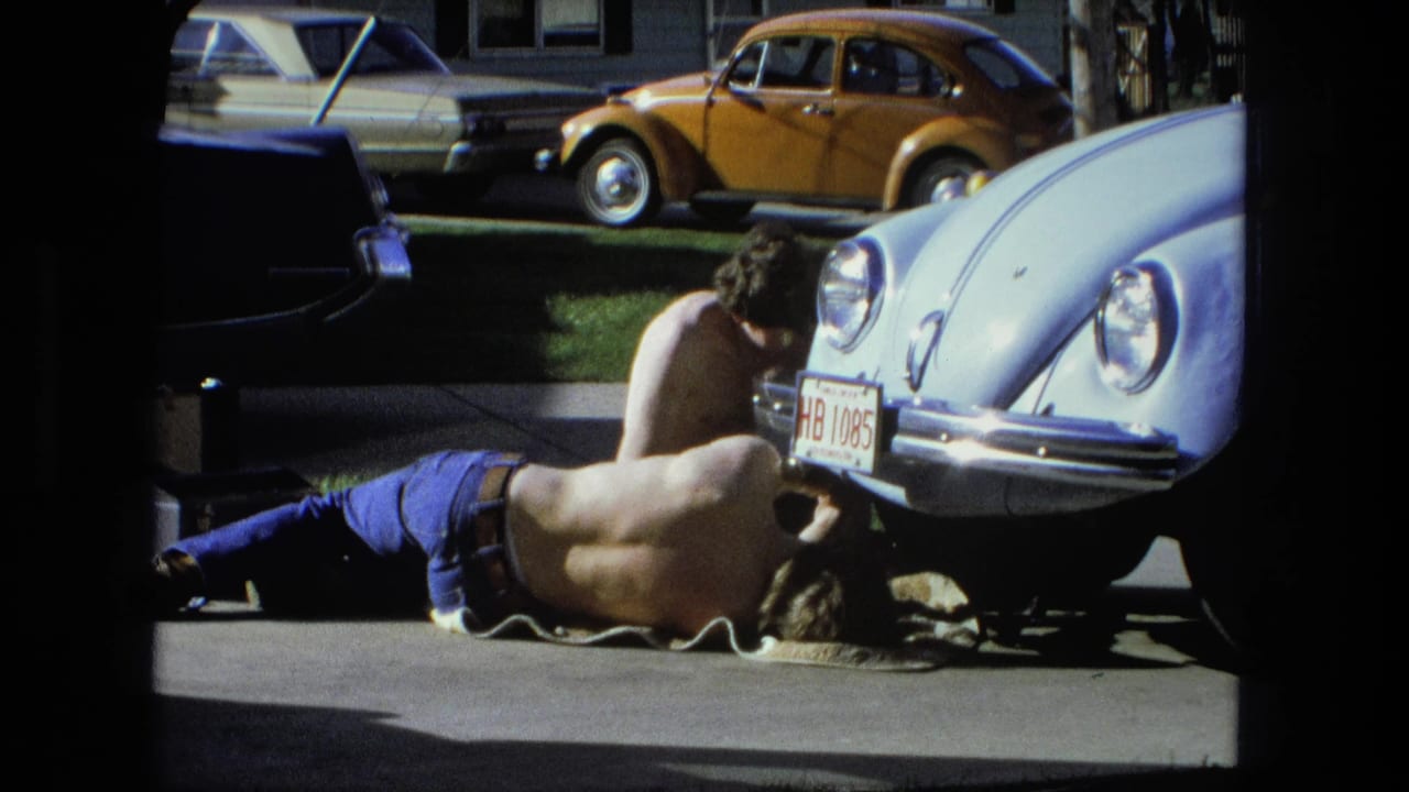 A man lies beneath the front of a light blue Beetle while another man works above him.