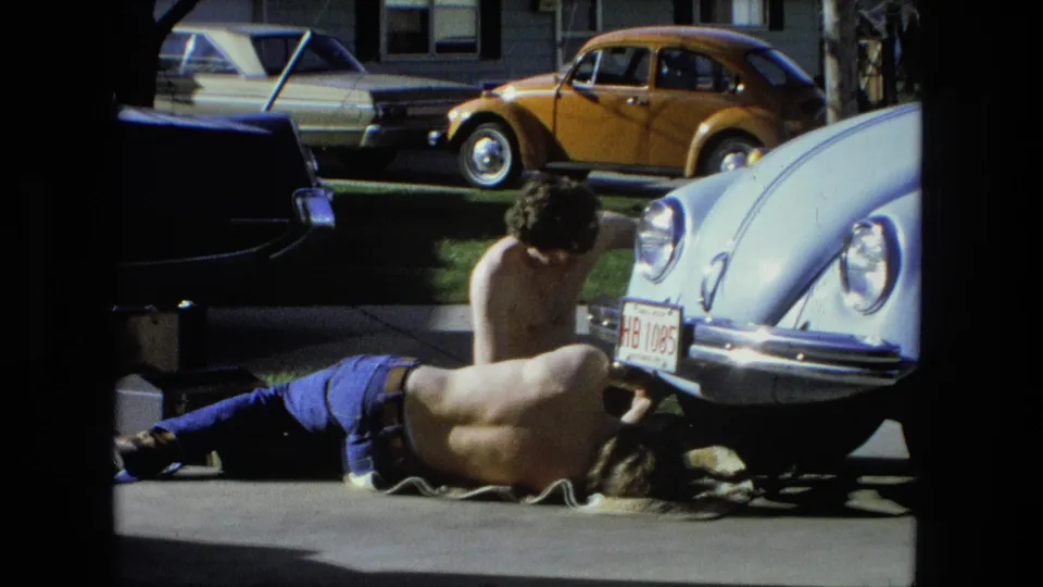 The home-movie frame shows two men repairing a light blue Volkswagen Beetle in a Fort Wayne driveway, with a yellow Beetle behind it.
