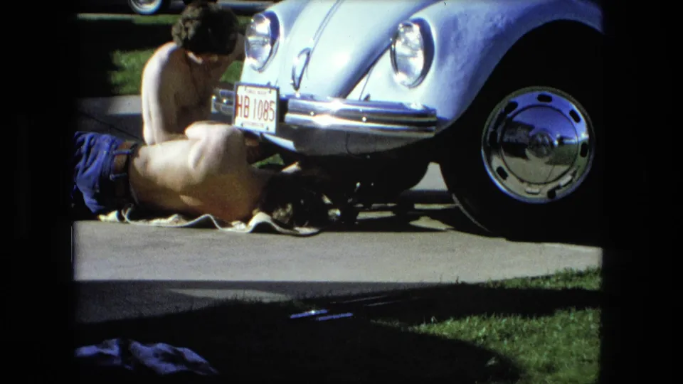 A closer 1976 Fort Wayne frame shows one man lying under a light blue Volkswagen Beetle while another sits near the bumper.