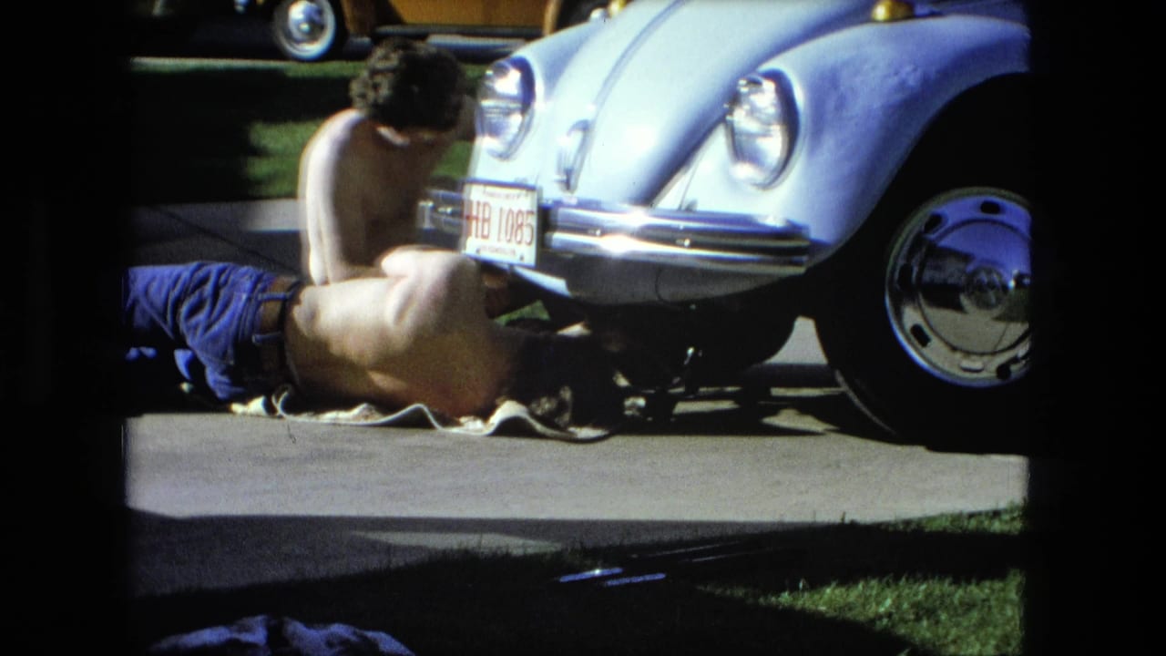 Two men work beside the front of a light blue Volkswagen Beetle in a sunny driveway.