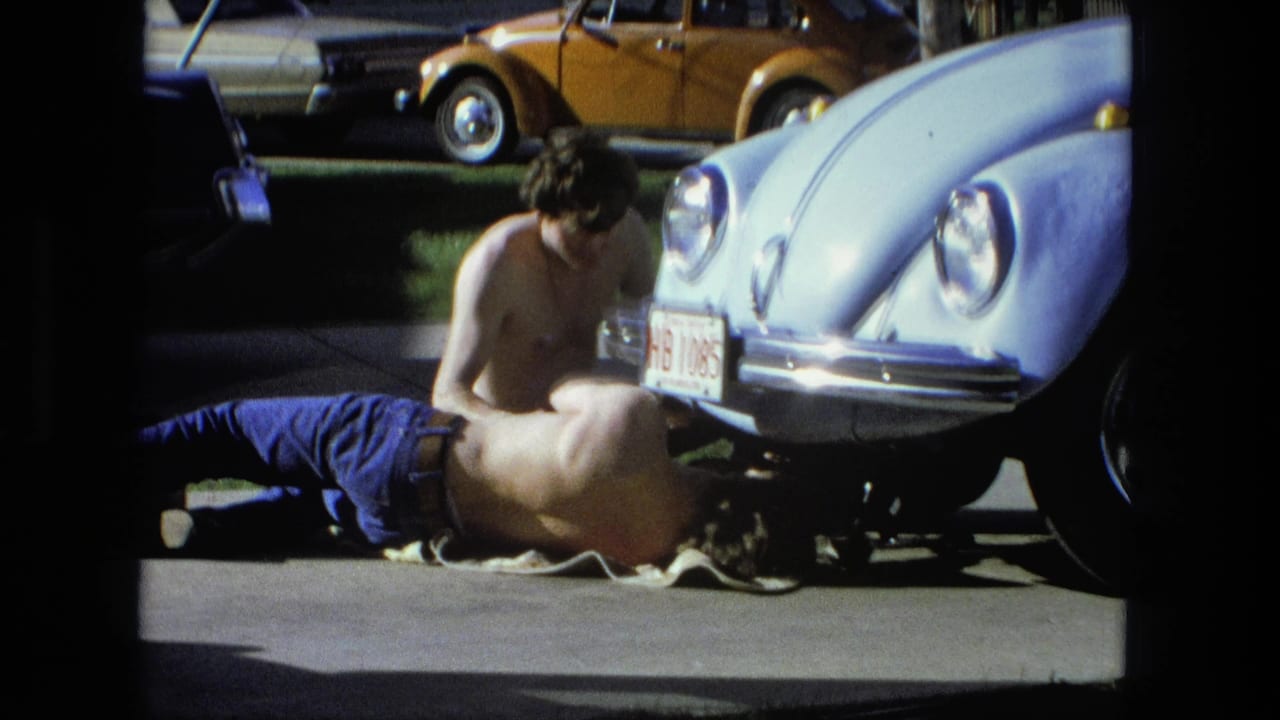 A shirtless man lies under a Beetle while another man leans over the front bumper.