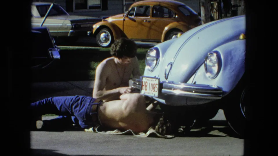 In the Fort Wayne 1976 home-movie scene, two men continue driveway repair work around the front of a light blue Beetle.