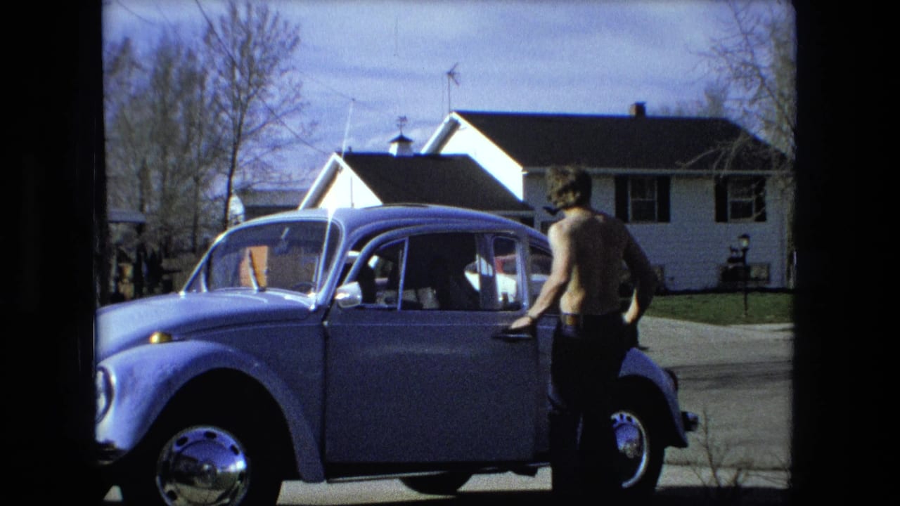Shirtless man stands beside a light blue compact car on a residential street.