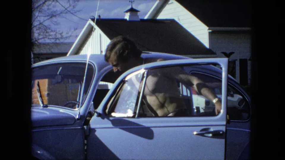 The man leans through the open driver-side door of the light blue car in a Fort Wayne residential setting from the 1976 8mm clip.