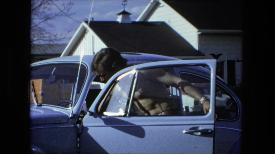 The 1976 Fort Wayne home-movie frame shows the man bent into the open car door, with the light blue vehicle filling most of the view.