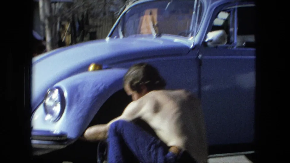At the end of the Fort Wayne 1976 clip, the shirtless man crouches near the front wheel area of the light blue car during the tire change.