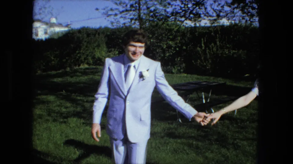 The groom in a light blue suit smiles toward the camera while holding the bride's hand in a Fort Wayne backyard.