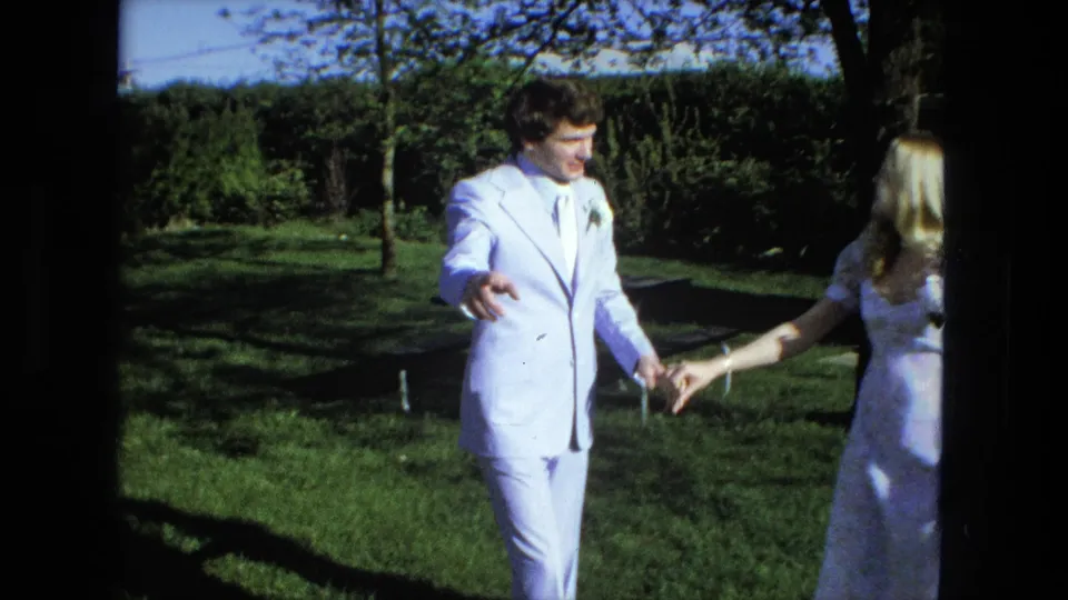The groom in a pale blue suit reaches toward the bride as they move apart across the sunny Fort Wayne lawn.