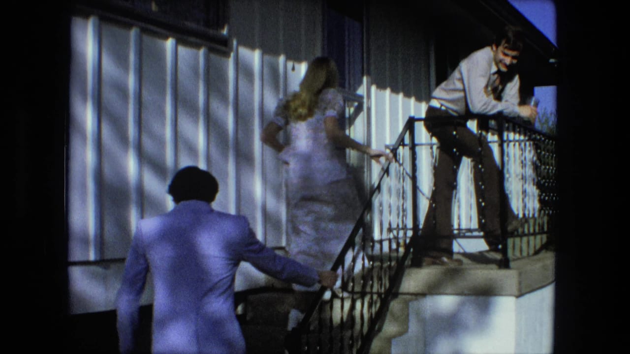 Blonde woman in a pale dress climbs porch stairs as a man in a periwinkle suit follows.