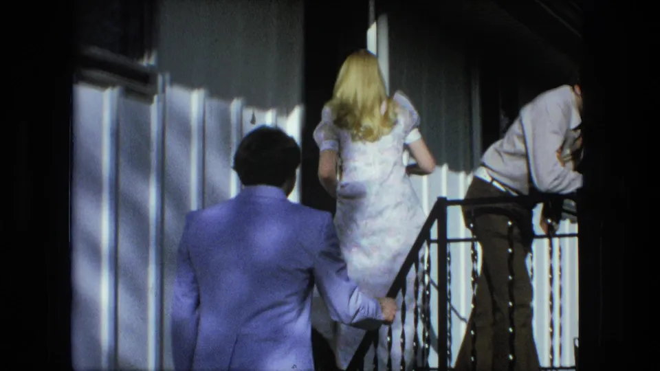 The couple continues up the porch steps in Fort Wayne, with a second young man leaning near the railing in the sunlit frame.