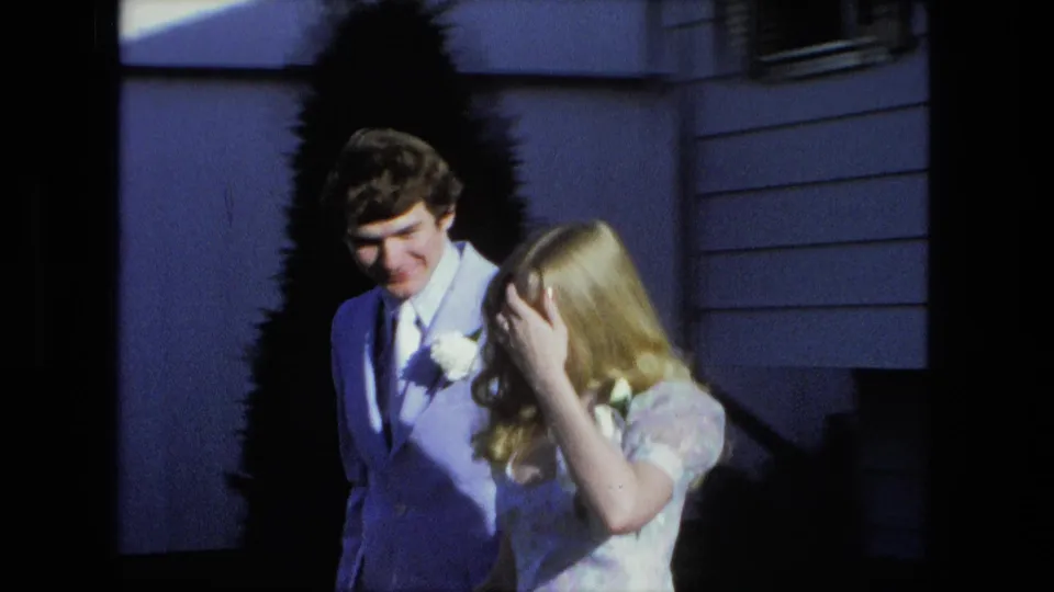 The couple stands together outside the house, with the man smiling and the blonde woman adjusting her hair in bright 8mm color.