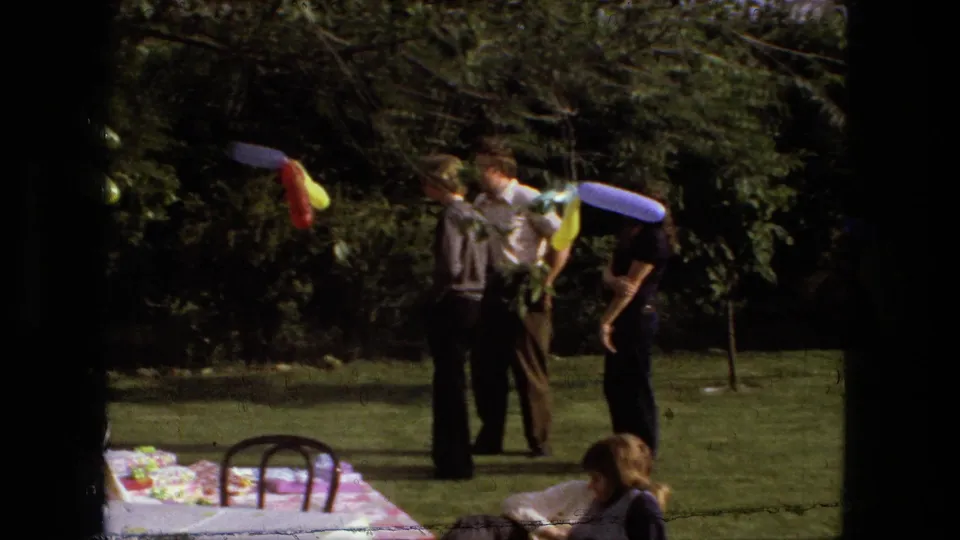 1976 Fort Wayne home-movie frame showing adults gathered on a backyard lawn with balloon decorations and a picnic table nearby.