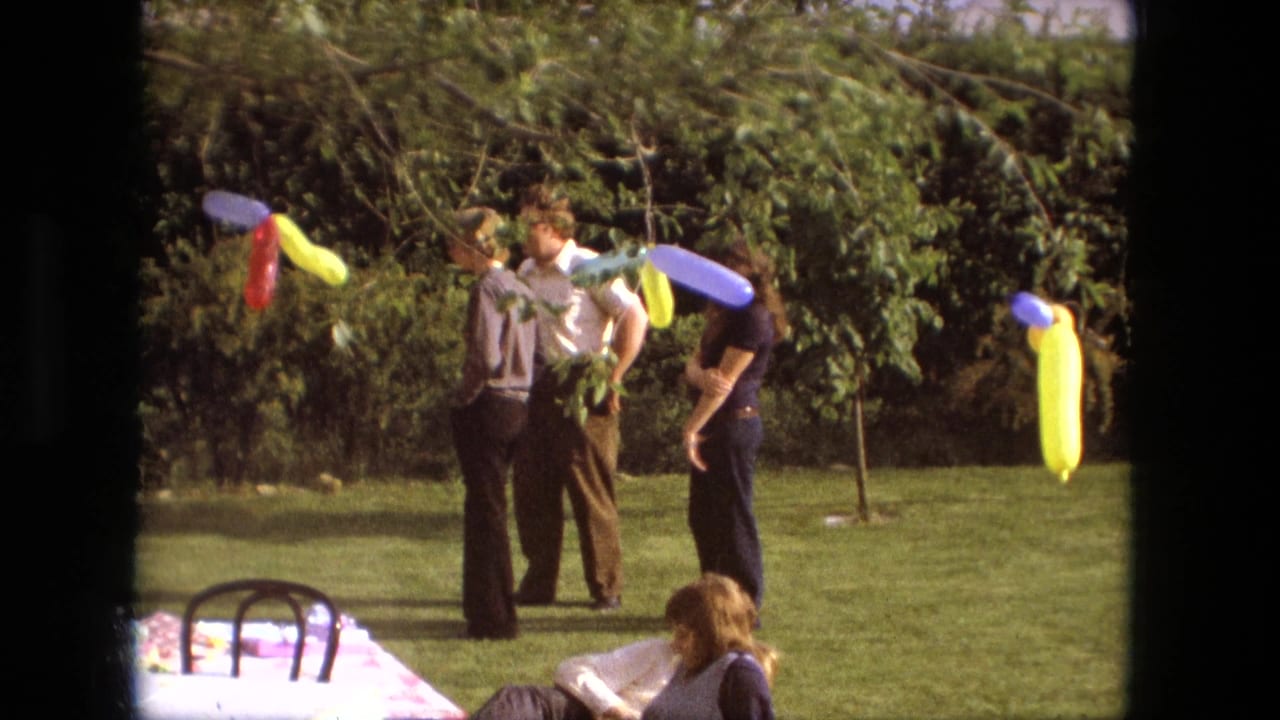 Fort Wayne, Indiana, 1976 frame of adults at an outdoor party, with bright twisted balloons and trees in the background.
