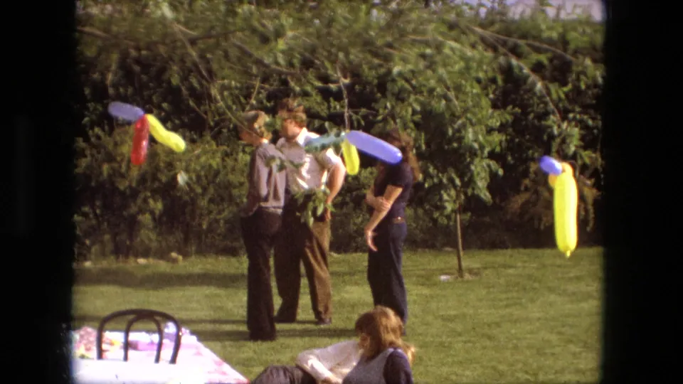 Fort Wayne, Indiana, 1976 frame of adults at an outdoor party, with bright twisted balloons and trees in the background.
