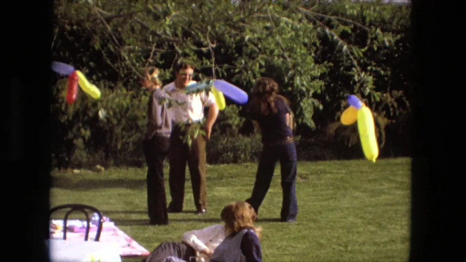 1976 backyard party frame showing adults in casual clothing near bright balloon decorations on a sunlit lawn in Fort Wayne.