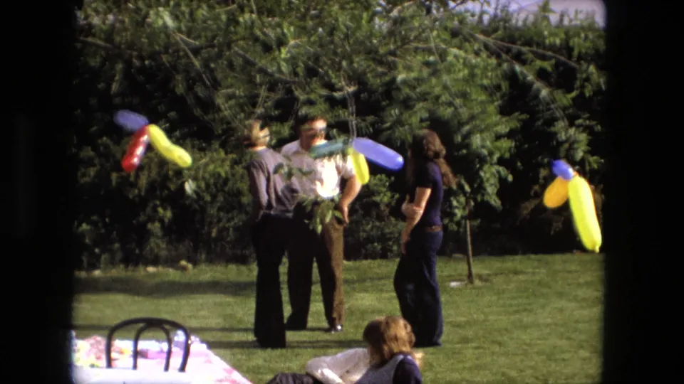 Fort Wayne 1976 home-movie still with adults talking on a backyard lawn, framed by trees and colorful balloon decorations.