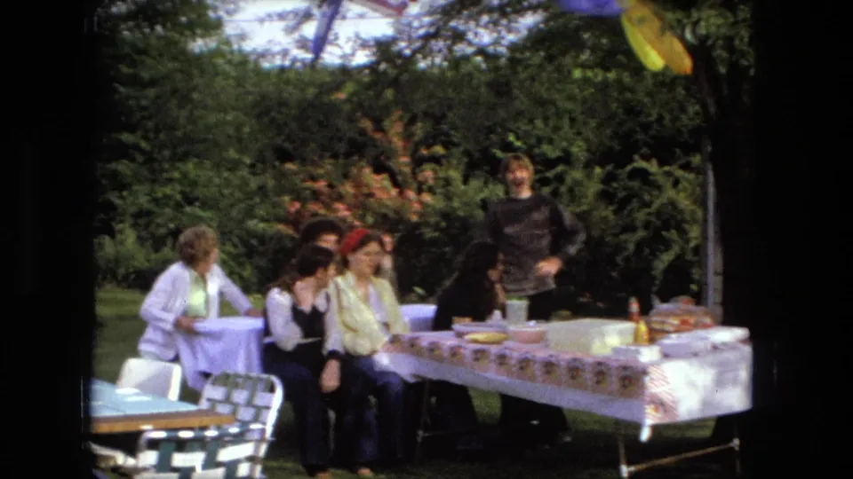 The 1976 Fort Wayne home movie shows adults seated at a backyard lunch table while another person stands beside them.