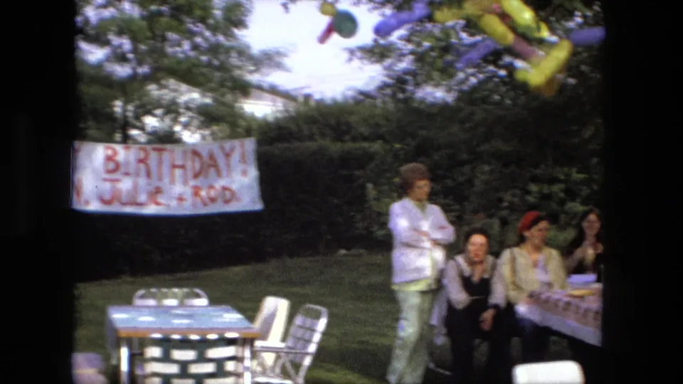 Fort Wayne 1976 home-movie frame with a handmade birthday banner, lawn chairs, picnic tables, balloons, and adult guests outdoors.