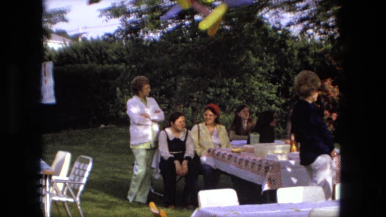 Adult guests sit and stand beside covered picnic tables in a backyard.