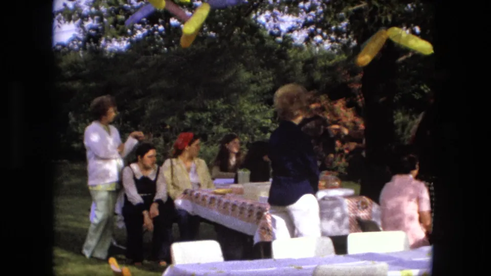 1976 Fort Wayne home-movie frame showing adults around a backyard party table under balloons and tree shade.