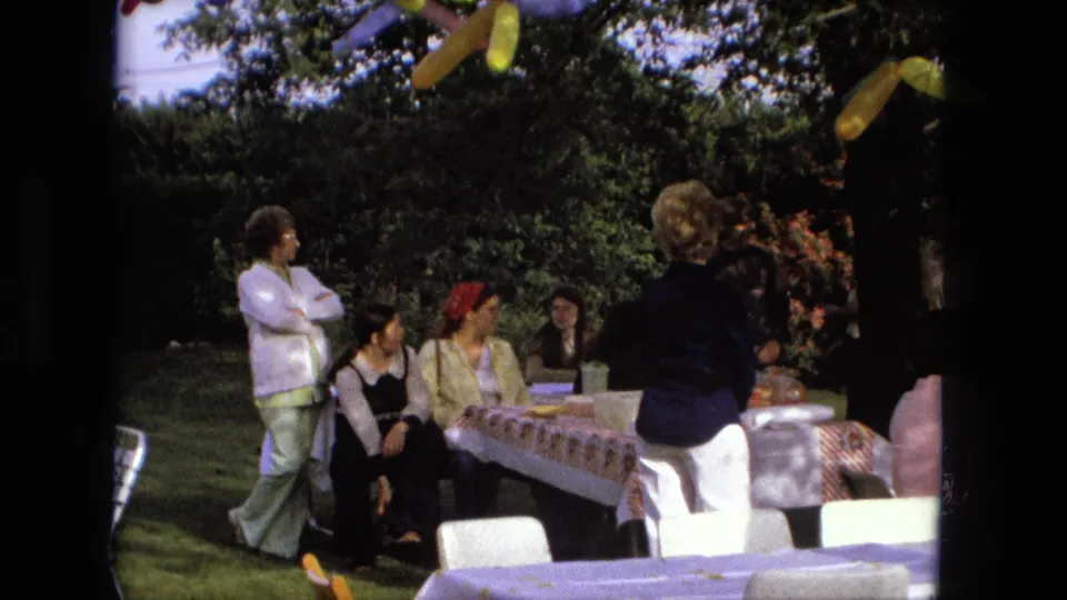Fort Wayne 1976 backyard birthday frame with seated and standing adults, covered picnic tables, balloons, hedges, and lawn chairs.