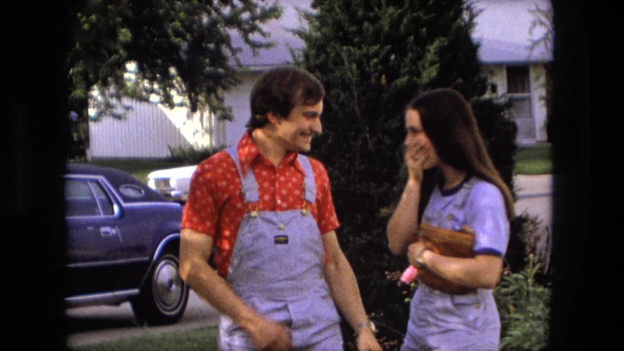 Young man and woman laugh together beside shrubs and a parked blue car.