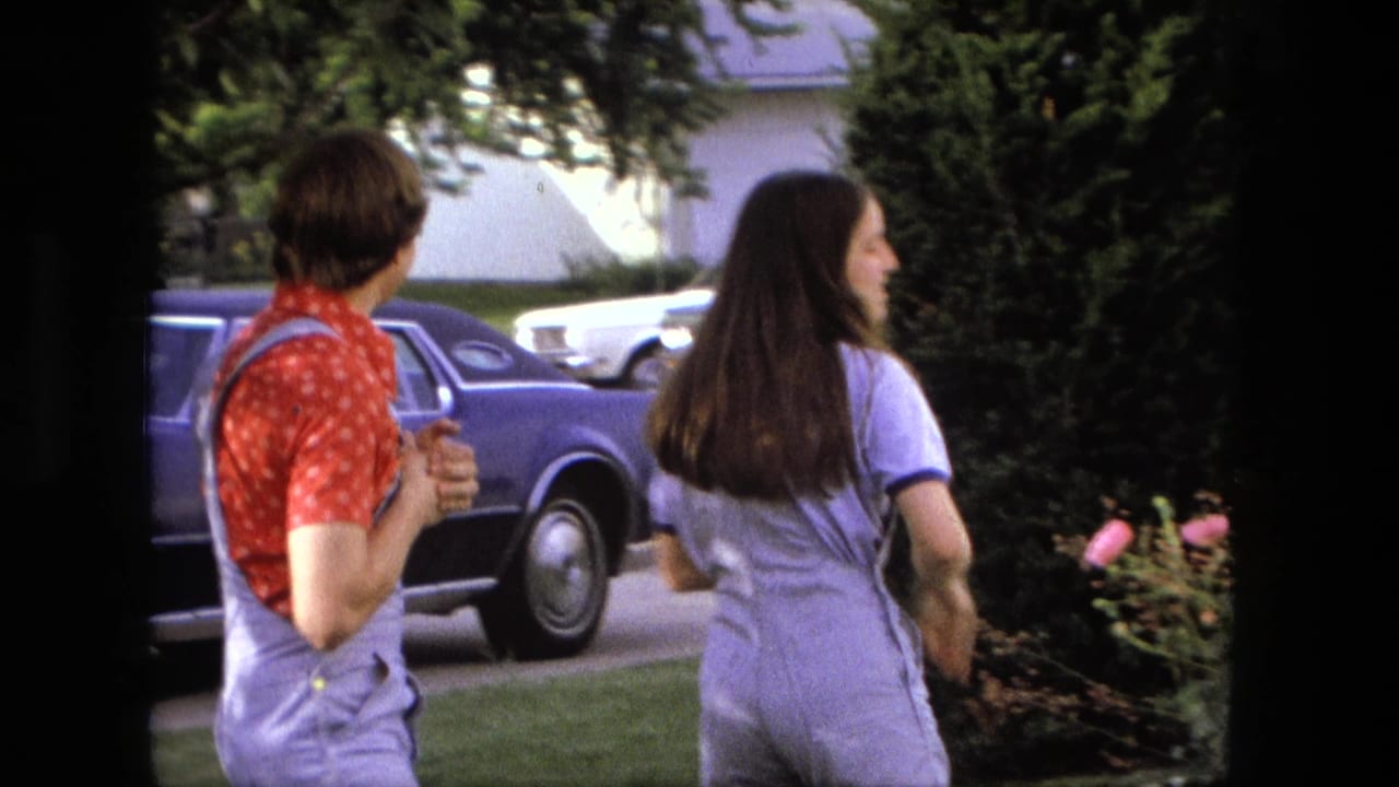 The woman turns away toward the yard while the young man watches near a parked blue car in Fort Wayne home-movie footage.