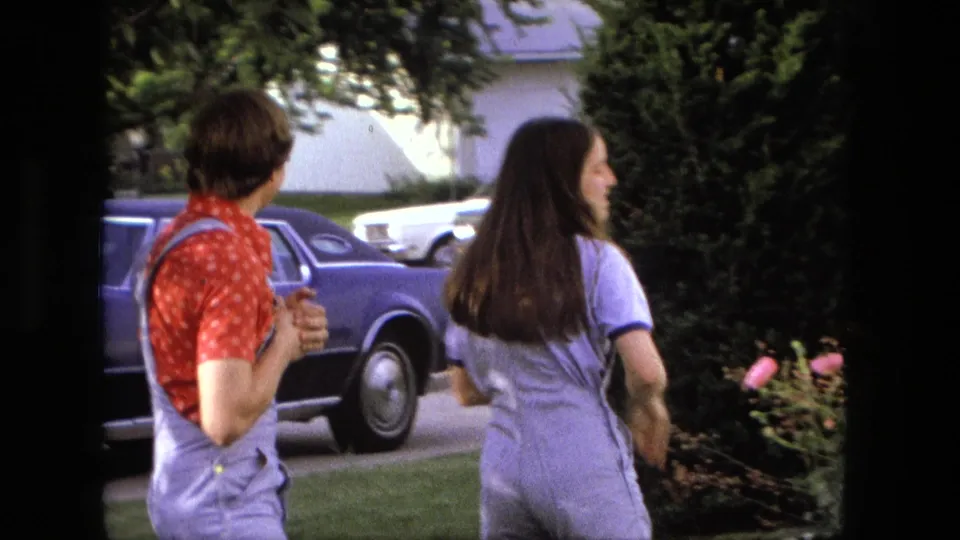 The woman turns away toward the yard while the young man watches near a parked blue car in Fort Wayne home-movie footage.