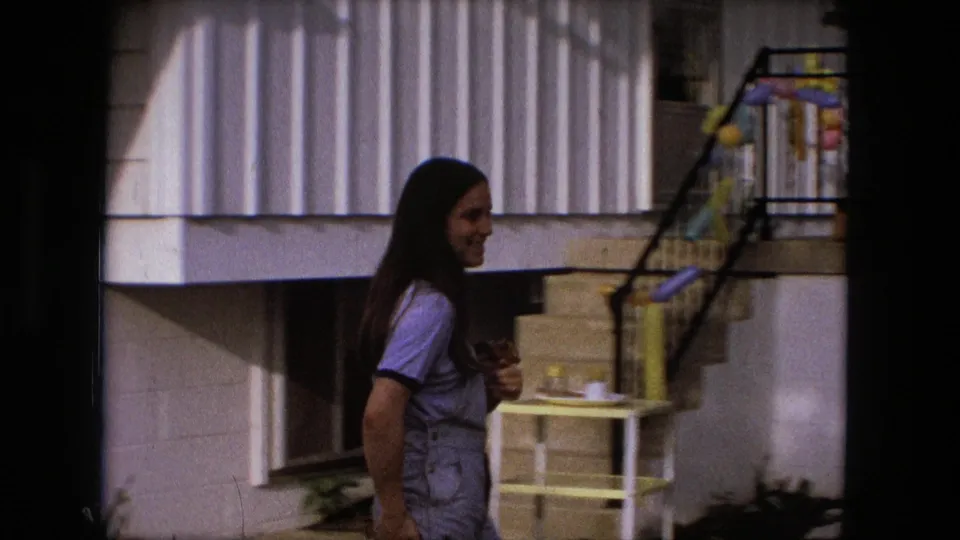 A young woman in overalls walks beside a house in Fort Wayne, with steps and colorful birthday decorations visible nearby.