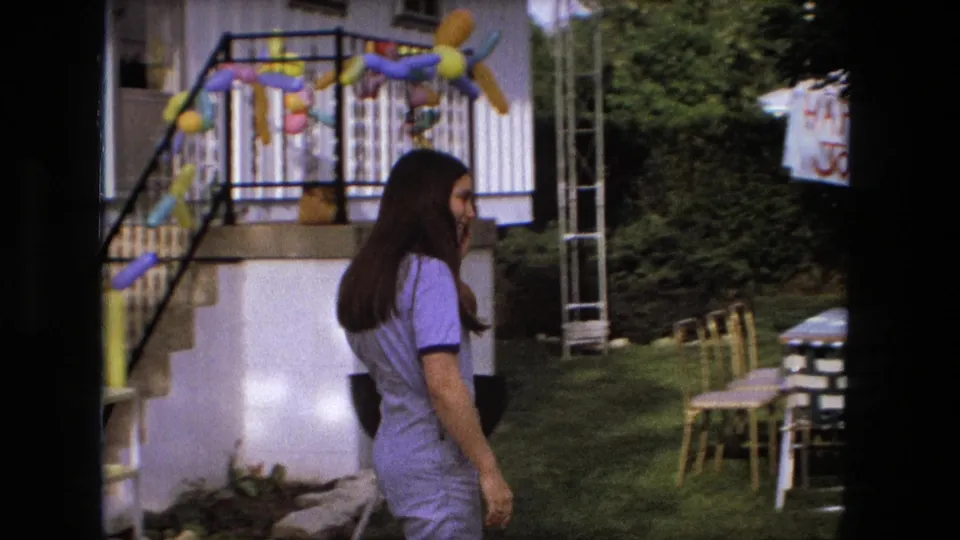 The woman approaches a backyard area with balloons, lawn chairs, and a visible birthday sign in 1976 Fort Wayne home-movie footage.