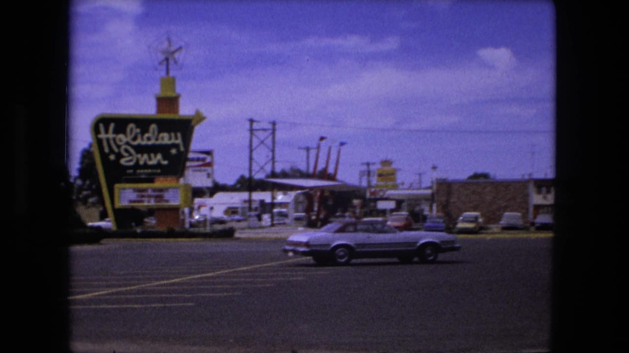 Holiday Inn sign beside a motel parking lot with a light convertible passing in front.