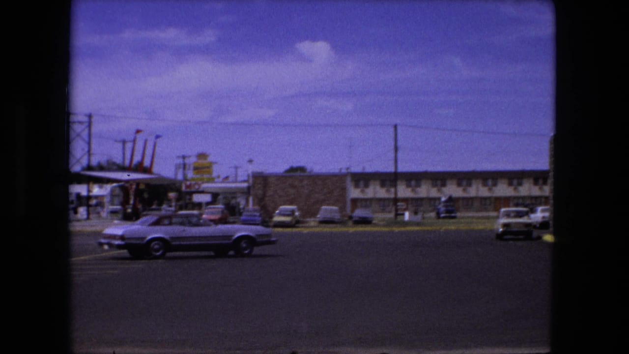 1976 Fort Wayne motel parking lot view with a light convertible moving past parked cars and low roadside buildings.