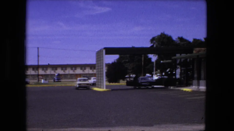 1976 Fort Wayne 8mm frame showing a motel parking lot, covered entry area, parked cars, and low building across the lot.