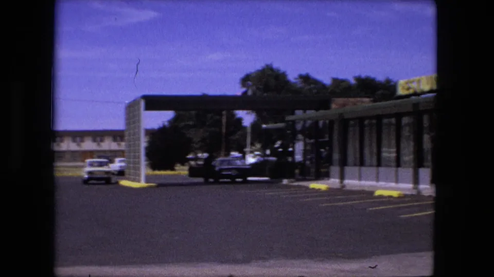 1976 Fort Wayne roadside restaurant exterior with a yellow sign, brick wall, covered walkway, yellow curb stops, and a few cars in a mostly empty parking lot.